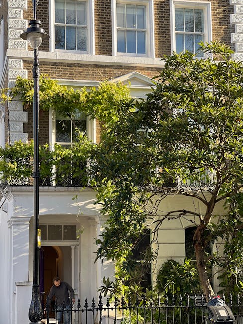 The image depicts the front facade of a period townhouse in Belgrave Square, featuring traditional brickwork with white window frames and decorative cornices. In the foreground, a well-maintained garden includes a large leafy tree with dense green foliage, partially obscuring the building. A black wrought iron fence borders the garden, and a classic streetlamp stands on the sidewalk beside the garden area. The garden bed beneath the tree is tidy, with some smaller plants and shrubs visible. A gardener is seen wearing dark clothing, engaged in outdoor maintenance or gardening tasks, near the white entrance door of the townhouse. The scene is illuminated by natural daylight, highlighting the textures of the wet soil in the garden beds, the trimmed grass, and the neatly kept plants. Overall, the setting emphasizes professional garden maintenance and landscaping work, reflecting careful attention to outdoor care typical of period townhouses in this area, as exemplified by Gardeners Belgravia.